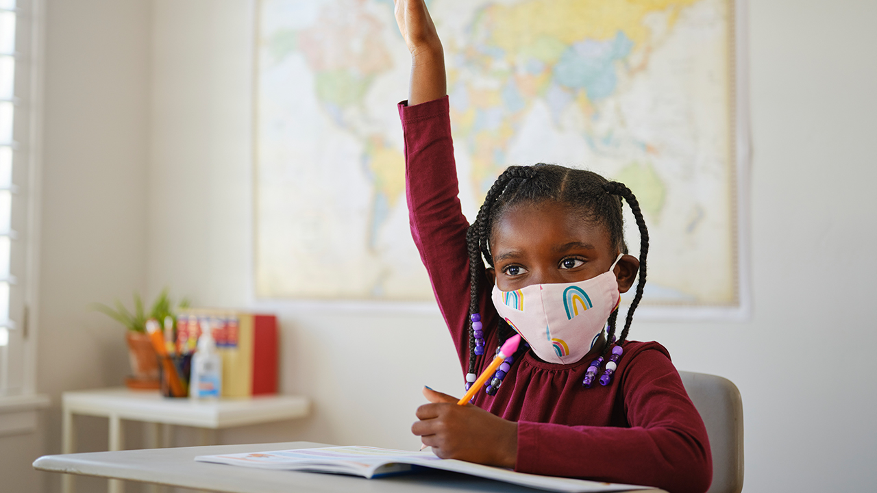 Foto de una niña con mascarilla puesta dentro de un salón de clases alzando la mano.